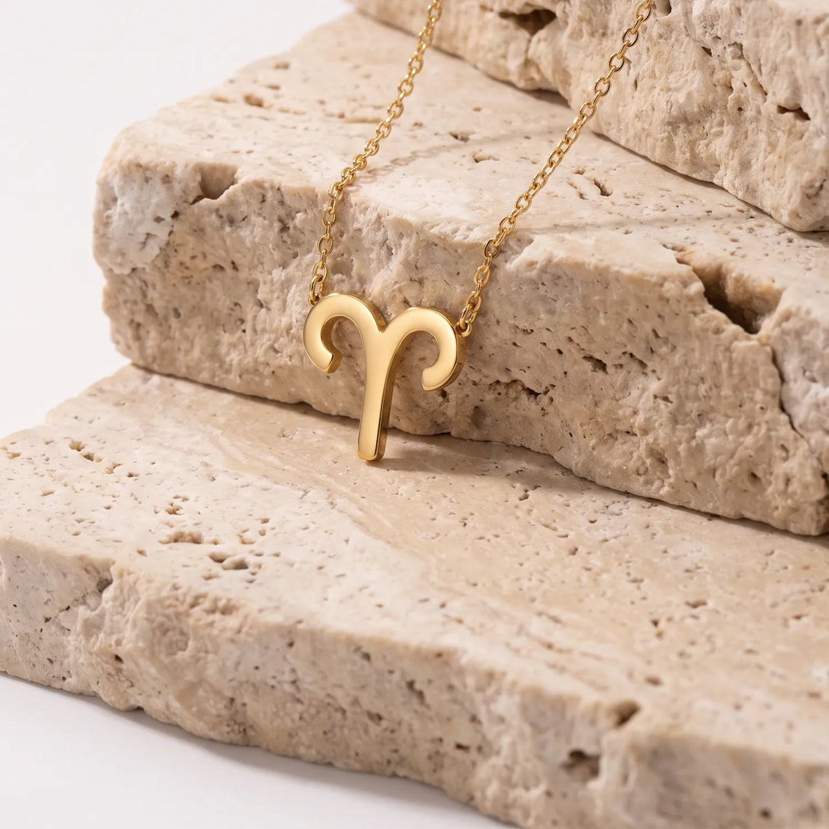 A gold necklace with a ram's head aries pendant, resting on stacked beige stone slabs against a clean white background.
