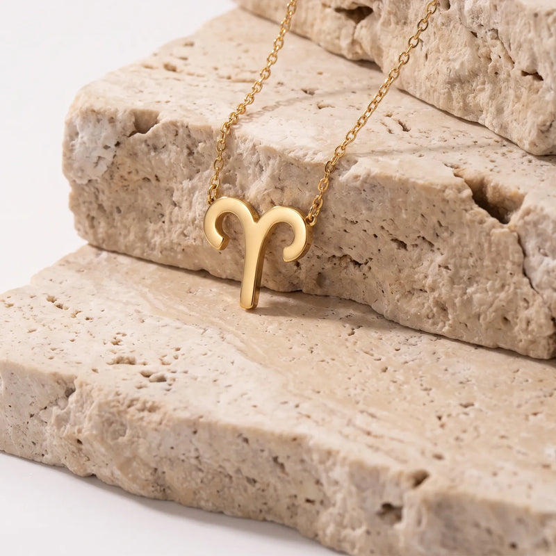 A gold necklace with a ram's head aries pendant, resting on stacked beige stone slabs against a clean white background.