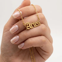 A gold nameplate necklace spelling Aries in a gothic font, worn on a delicate gold chain and held by a hand with manicured nails against a clean white background.