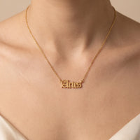 A gold nameplate necklace spelling 'Aries' in a gothic script worn on a model's neck, shown in a close-up shot against a warm beige background.