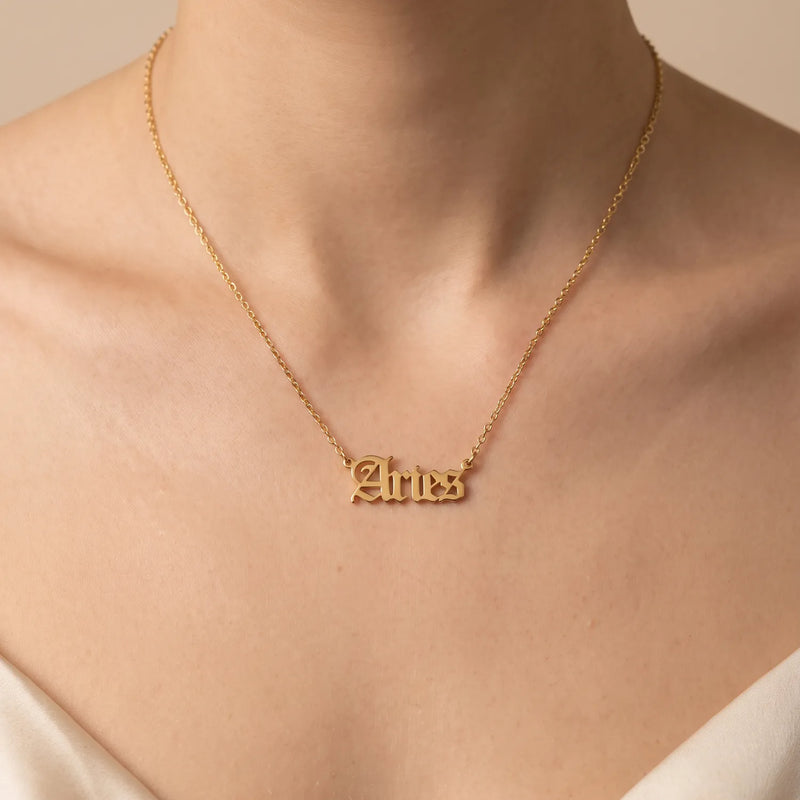 A gold nameplate necklace spelling 'Aries' in a gothic script worn on a model's neck, shown in a close-up shot against a warm beige background.