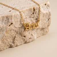 A gold nameplate necklace spelling 'Aries' in a celtic-style script, resting on a textured beige stone prop against a warm beige background.