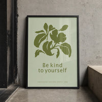 Minimalist botanical art print with olive-green leaves and the words “Be kind to yourself” on a pale green background, displayed in a black wood frame.