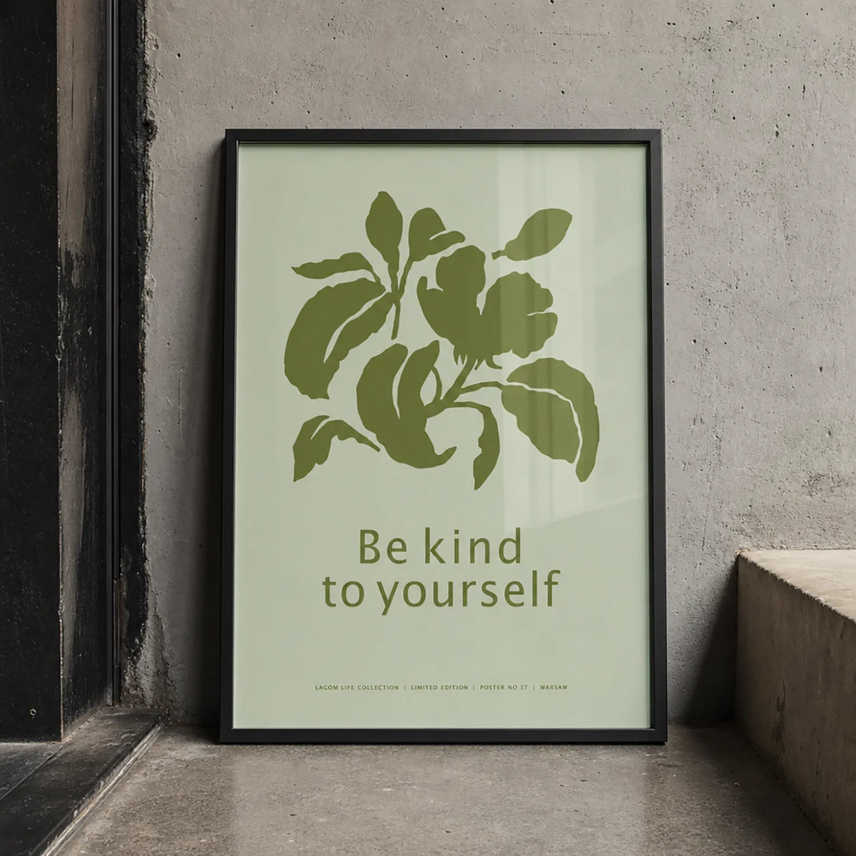 Minimalist botanical art print with olive-green leaves and the words “Be kind to yourself” on a pale green background, displayed in a black wood frame.