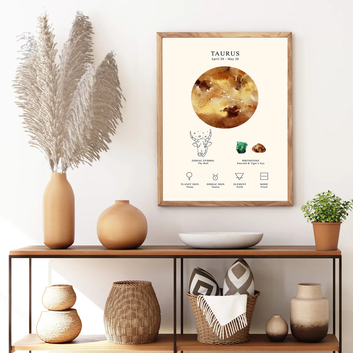 A framed Taurus astrology print hangs on a white wall above a wooden console table styled with vases, a bowl, and a small plant. The print features a cream background with a large circular astrological symbol, the word 'TAURUS,' and small line illustrations and text.