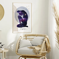 A framed wall art print featuring a seated Capricorn figure in a purple galaxy pattern against a pale blue moon, displayed above a rattan armchair in a styled room.