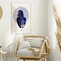 A framed wall art print hangs above a light wood rattan armchair in a styled interior. The artwork shows an abstract human silhouette filled with a galaxy-like blue, purple, and gold pattern on a white background with gold geometric lines.