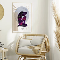 A framed wall art print featuring a stylized seated figure in a purple galaxy pattern against a pale blue circle, hung above a woven rattan armchair in a bright, neutral-toned room.