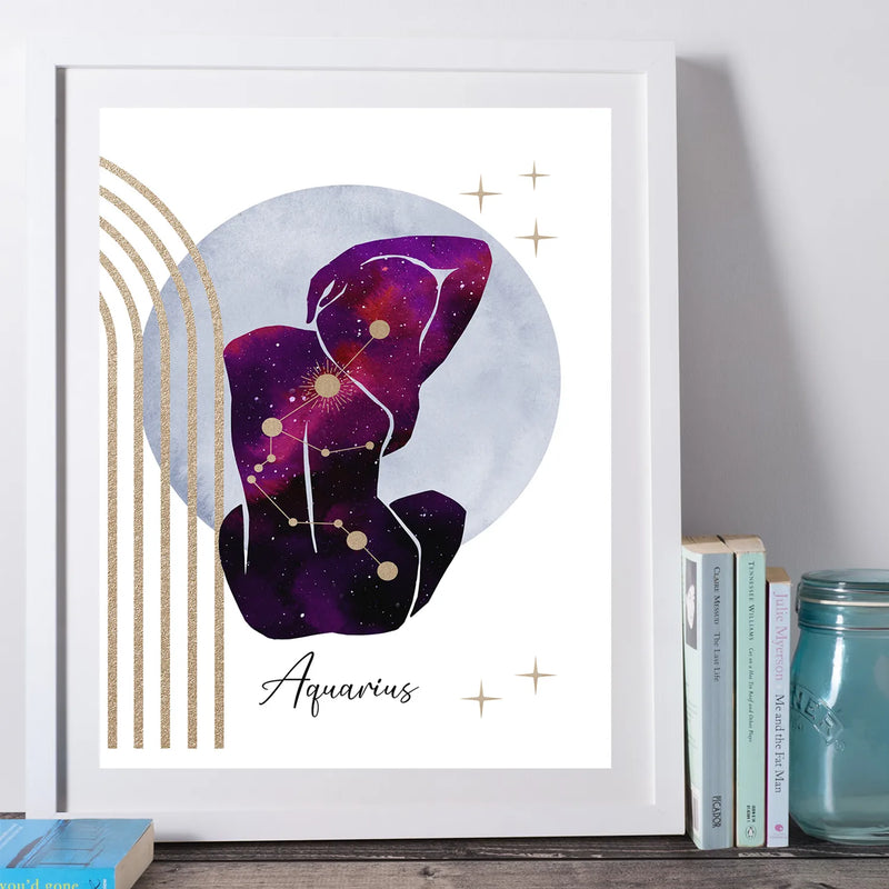A framed art print featuring a purple and magenta watercolor galaxy constellation design labeled 'Aquarius' with gold accents, styled leaning against a wall next to books and a glass jar.