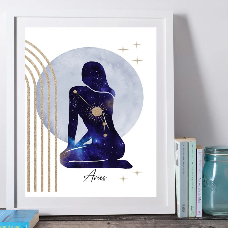 A framed art print featuring a stylized Aries zodiac silhouette with a galaxy and sun design, set against a large moon and gold arches. The print is in a white frame and styled on a wooden surface next to books and a glass jar.