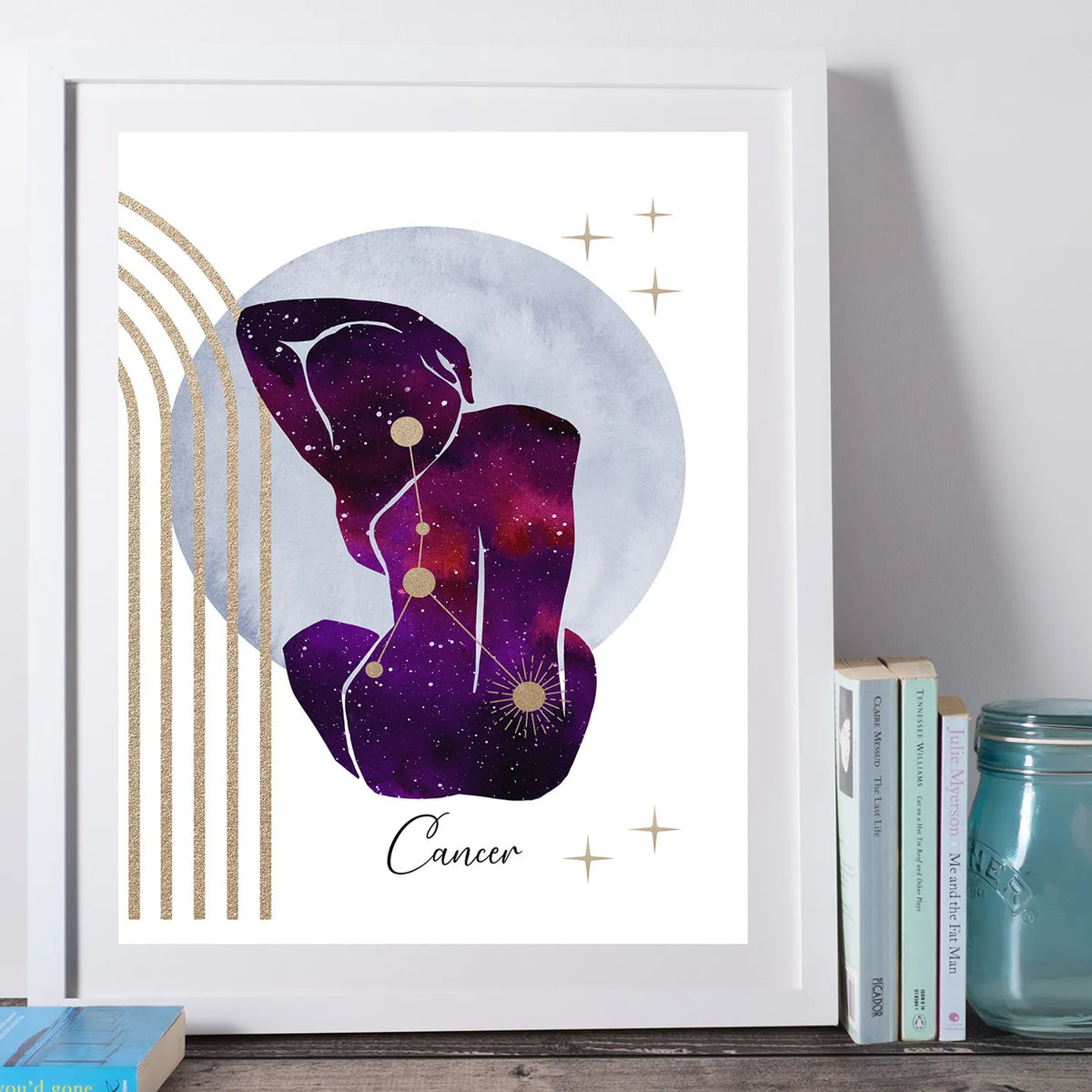 A framed art print featuring a purple and pink galaxy-style Cancer zodiac sign illustration with gold stars and stripes, displayed leaning against a wall on a wooden surface next to books and a glass jar.
