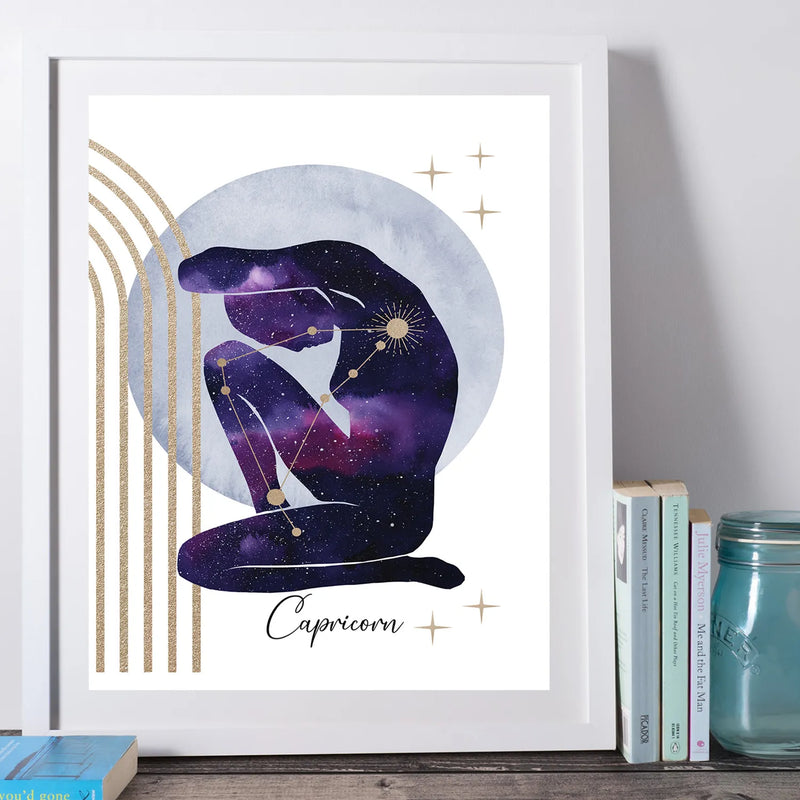 A framed Capricorn art print featuring a purple and blue galaxy silhouette of a seated figure with gold constellation lines and text, displayed leaning against a wall on a wooden surface next to books and a mason jar.