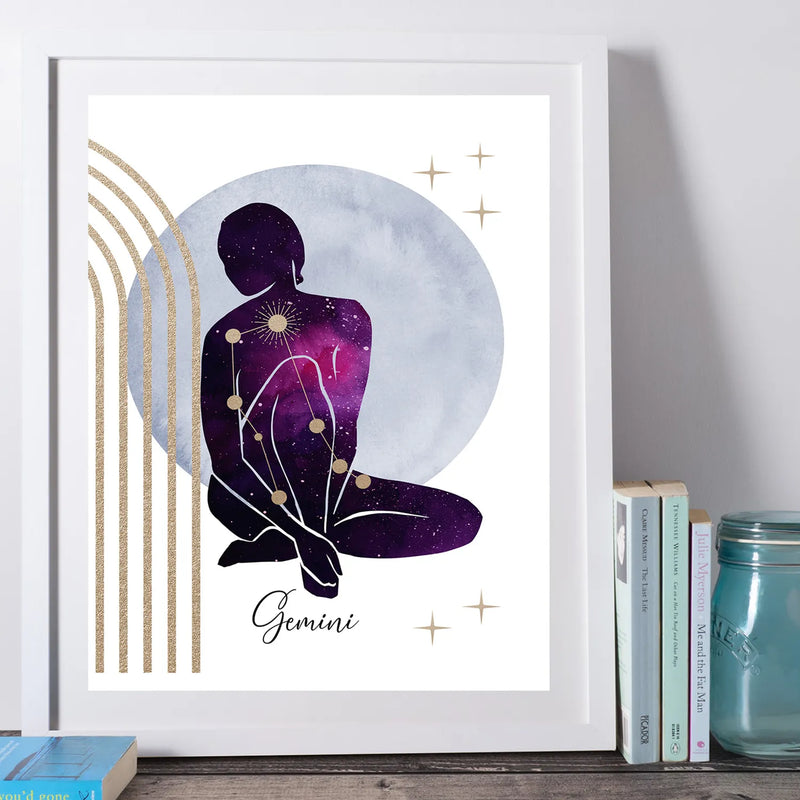 A framed art print featuring a Gemini zodiac sign illustration, styled on a wooden surface next to books and a glass jar. The print shows a seated Gemini silhouette in a purple and black galaxy pattern against a light blue-gray moon, with gold accents and the word 'Gemini' at the bottom.
