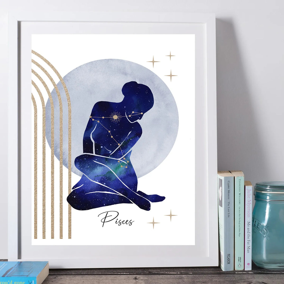 A framed art print featuring a Pisces zodiac sign illustration with a seated figure in a galaxy pattern, set against a large gray-blue circle and gold stars, styled on a shelf next to books and a mason jar.