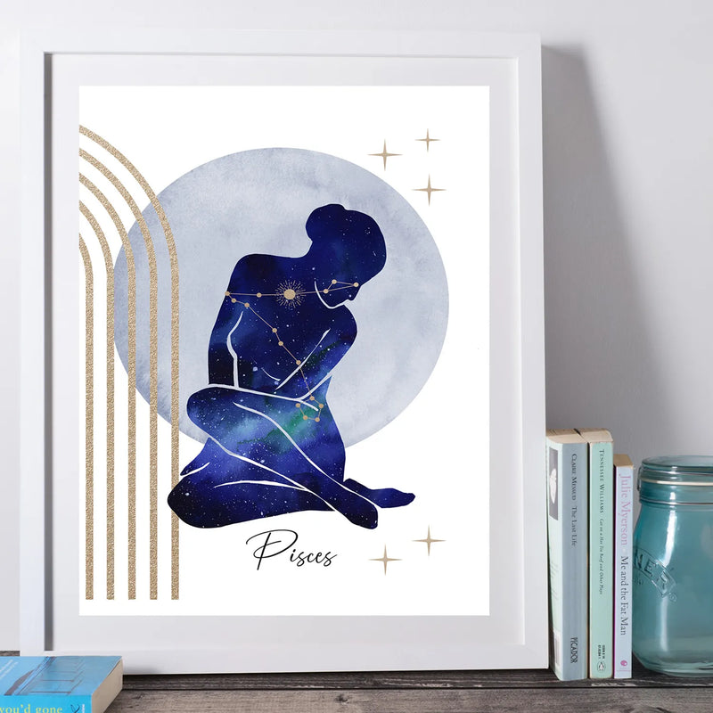 A framed art print featuring a Pisces zodiac sign illustration with a seated figure in a galaxy pattern, set against a large gray-blue circle and gold stars, styled on a shelf next to books and a mason jar.