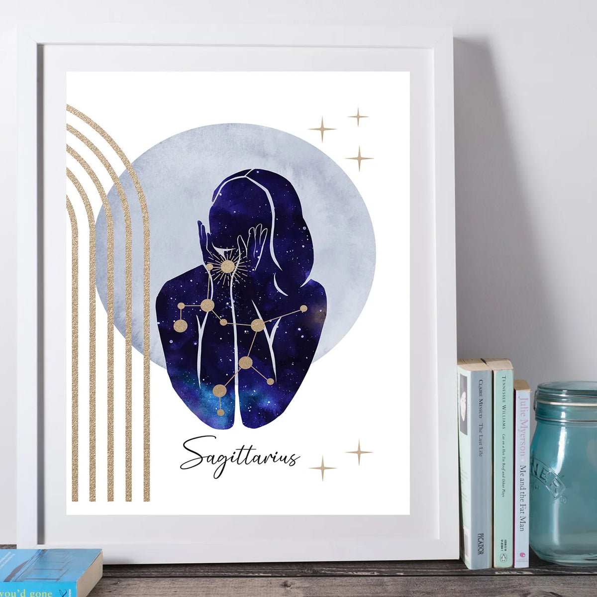 A framed art print featuring a stylized Sagittarius zodiac illustration with a dark blue watercolor figure, gold stars, and gold vertical stripes on a white background, leaning against a wall beside books and a mason jar.