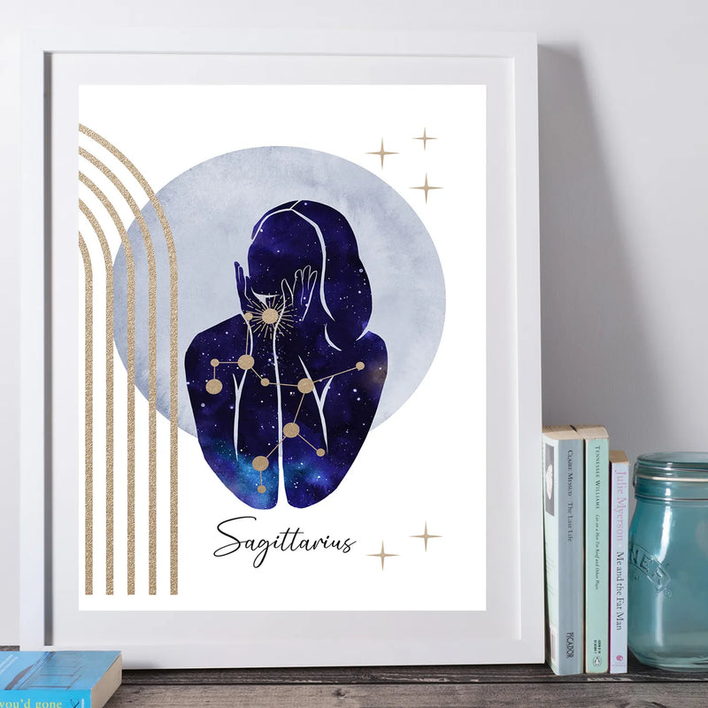 A framed art print featuring a stylized Sagittarius zodiac illustration with a dark blue watercolor figure, gold stars, and gold vertical stripes on a white background, leaning against a wall beside books and a mason jar.