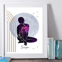 A framed art print featuring a seated Scorpio zodiac figure in a purple and pink galaxy pattern, set against a large pale blue circle with gold stars and arches. The print is housed in a white frame and styled on a wooden surface next to books and a glass jar.