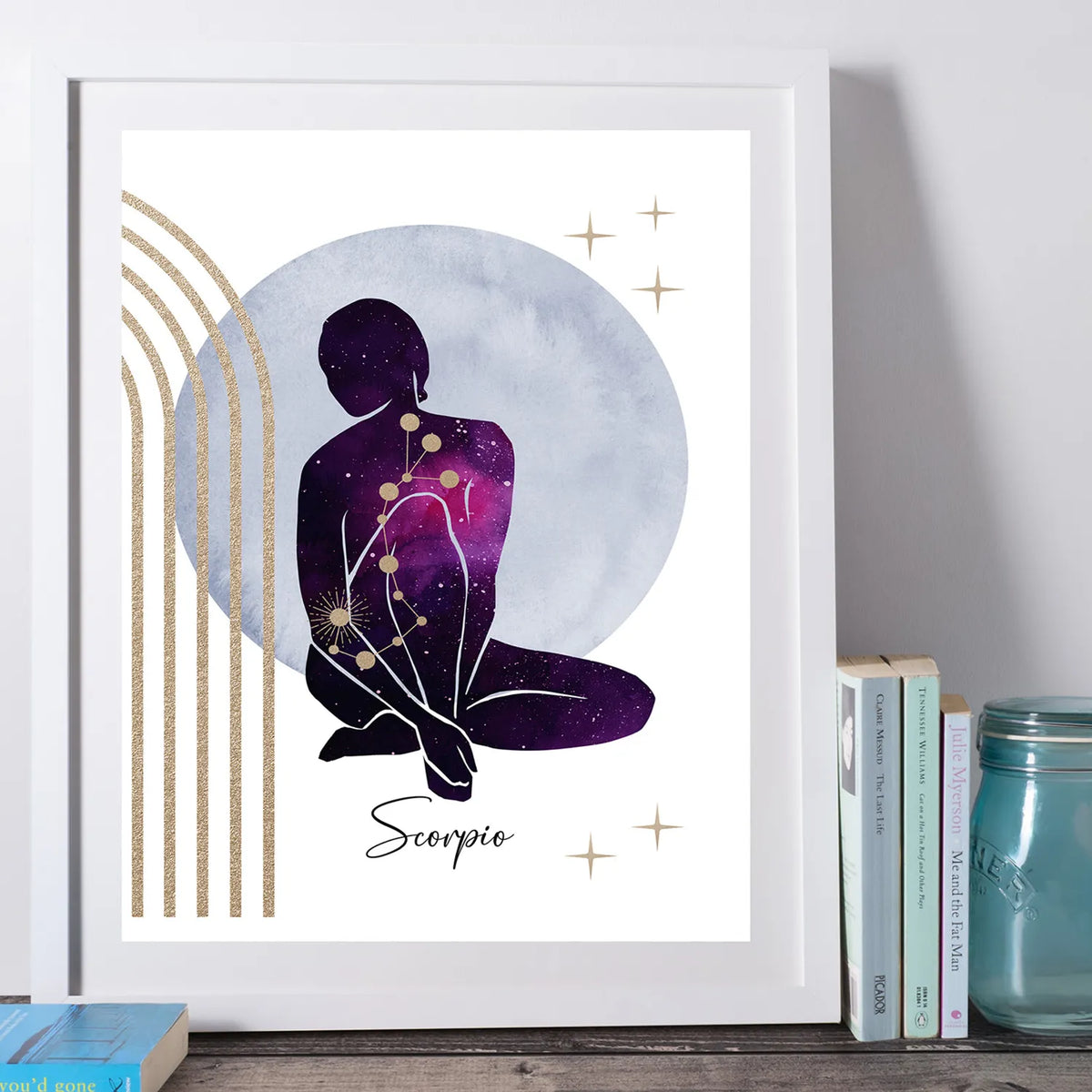 A framed art print featuring a seated Scorpio zodiac figure in a purple and pink galaxy pattern, set against a large pale blue circle with gold stars and arches. The print is housed in a white frame and styled on a wooden surface next to books and a glass jar.