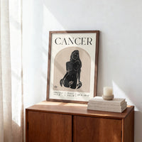 A framed art print featuring a black ink illustration of a female nude under the word 'CANCER' on a cream background, leaning against a white wall on a wooden cabinet next to a stack of books and a candle.