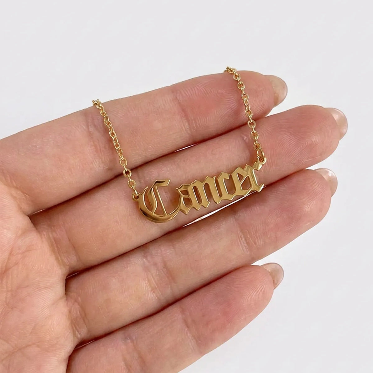 A gold nameplate necklace spelling 'Cancer' in a gothic font, held on a person's fingers against a plain white background.