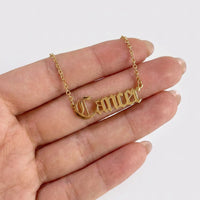 A gold nameplate necklace spelling 'Cancer' in a gothic font, held on a person's fingers against a plain white background.
