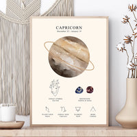 A framed Capricorn zodiac art print displayed on a wooden surface next to a macrame wall hanging and a vase with dried flowers. The print features a watercolor-style zodiac symbol, astrological details, and text for Capricorn.