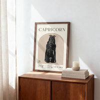 A framed Capricorn art print with a black illustration of a horned figure on a cream background, styled on a wooden sideboard next to a candle and books in a sunlit room.