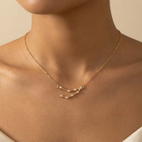 A close-up of a model's neck and upper chest wearing a delicate gold chain necklace with a capricorn constellation pendant set with tiny clear stones, against a soft beige background.