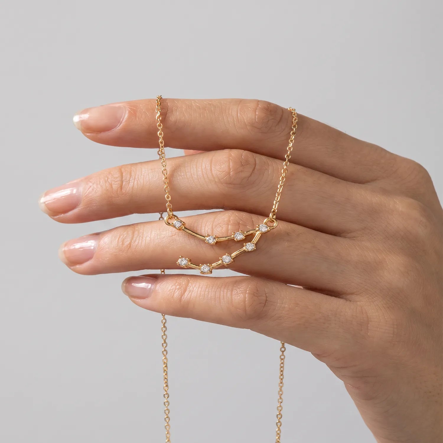 A gold necklace with a delicate, capricorn constellation set with small clear stones, held by a hand against a neutral light gray background.