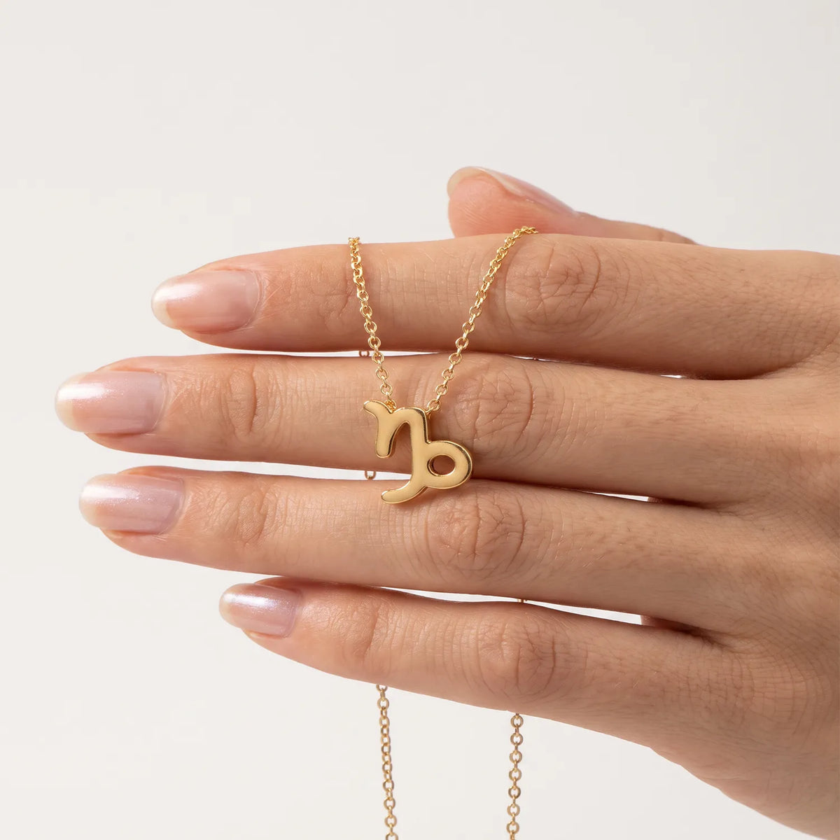 A gold necklace with a capricorn zodiac sign pendant held on a person's fingers against a clean, light off-white background.