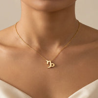 A close-up of a model's neck and upper chest wearing a delicate gold chain necklace with a capricorn zodiac sign pendant.