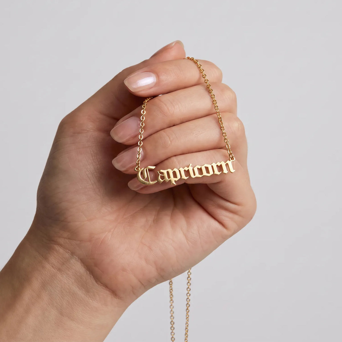 A hand holding a gold chain necklace with a 'Capricorn' nameplate pendant in a gothic script against a light gray background.