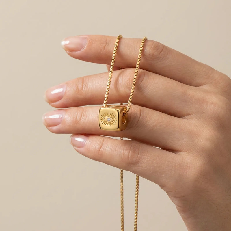 A gold necklace with a cube pendant featuring a evil eye design and a small central gemstone, held on a person's fingers against a warm beige background.