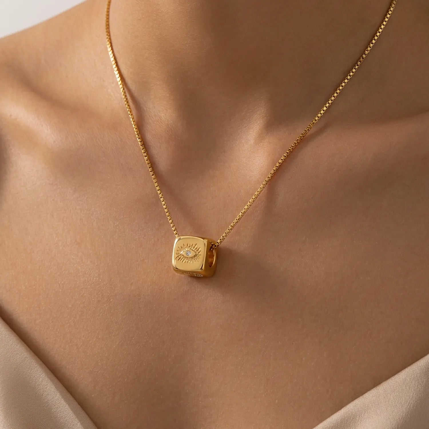 A close-up of a gold necklace with a cube pendant featuring an eye design, worn on a model's neck.