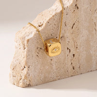 A gold cube pendant necklace with an evil eye design and a half moon , draped over a textured beige stone block on a light beige background.