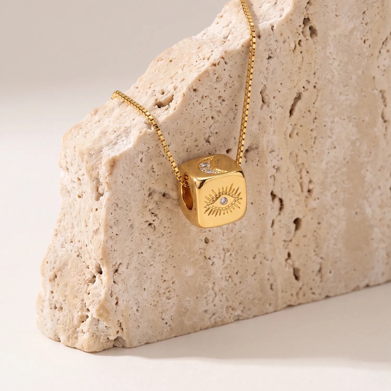 A gold cube pendant necklace with an evil eye design and a half moon , draped over a textured beige stone block on a light beige background.