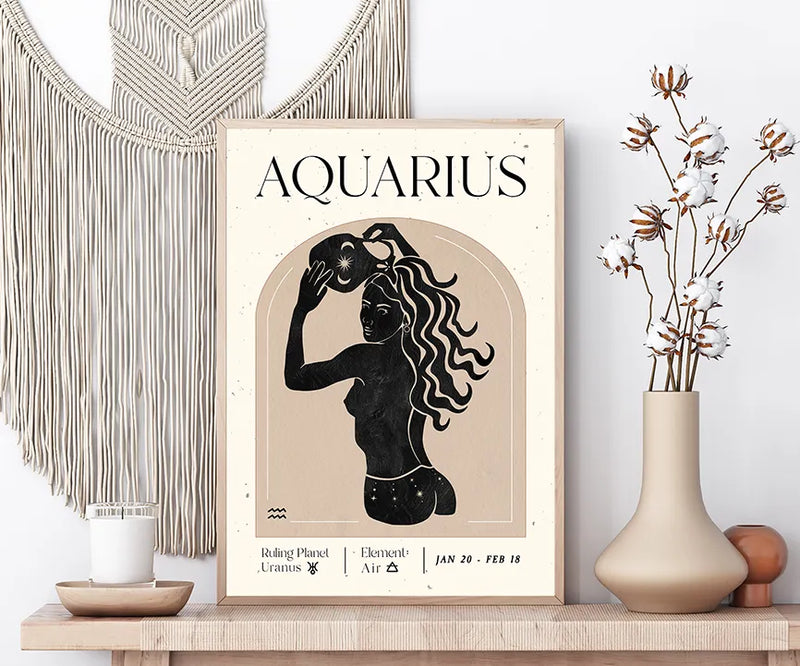 A framed art print featuring a black silhouette of an Aquarius zodiac figure on a beige background, styled on a wooden shelf next to a macrame wall hanging and a vase with dried flowers.