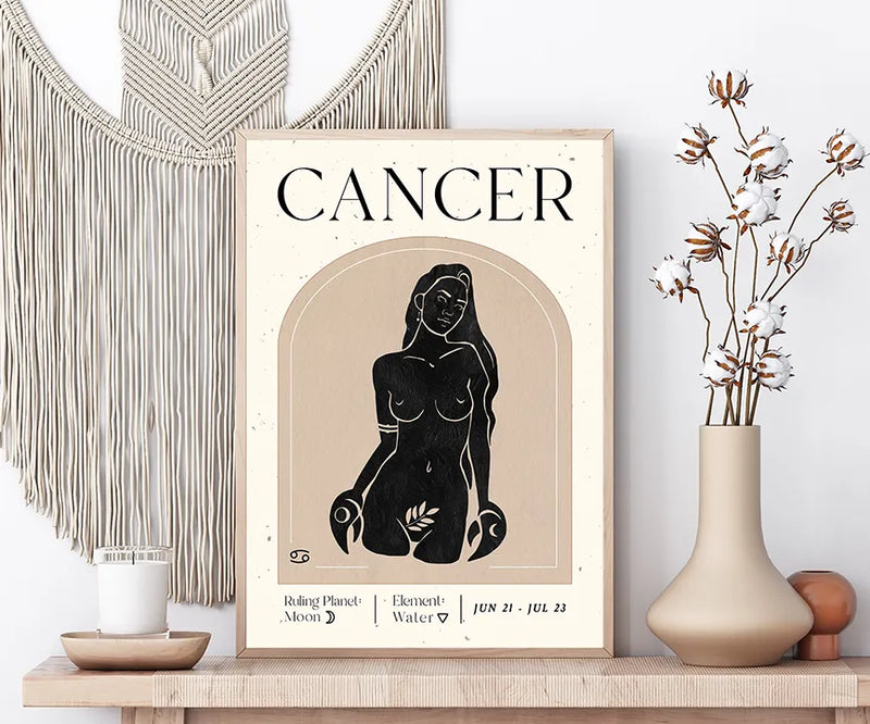 A framed art print featuring a vintage-style Cancer zodiac poster with a black silhouette of a woman on a beige background, styled on a wooden shelf next to a macrame wall hanging, a candle, and a vase with dried flowers.