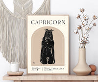 A framed Capricorn art print displayed on a wooden shelf in a styled room setting, flanked by a macrame wall hanging, a candle, and a vase with dried cotton stems.