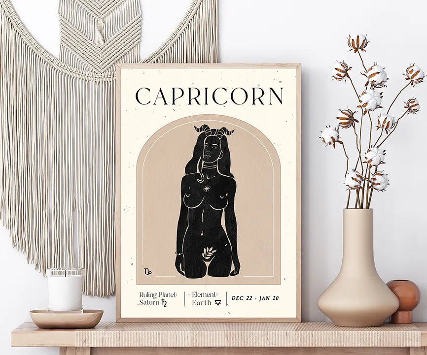 A framed Capricorn art print displayed on a wooden shelf in a styled room setting, flanked by a macrame wall hanging, a candle, and a vase with dried cotton stems.