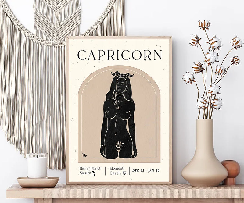 A framed Capricorn art print displayed on a wooden shelf in a styled room setting, flanked by a macrame wall hanging, a candle, and a vase with dried cotton stems.