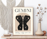 A framed Gemini astrology art print displayed on a wooden shelf in a styled room setting, flanked by a macrame wall hanging, a candle, and a vase with dried cotton stems.