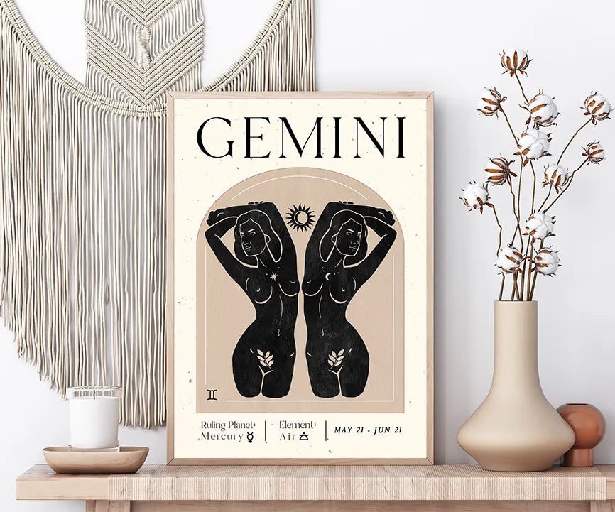 A framed Gemini astrology art print displayed on a wooden shelf in a styled room setting, flanked by a macrame wall hanging, a candle, and a vase with dried cotton stems.