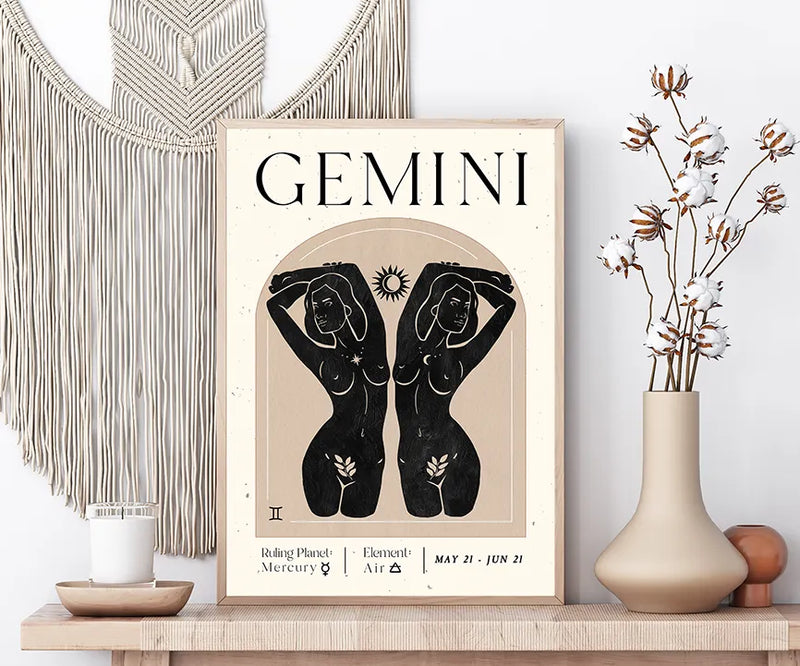 A framed Gemini astrology art print displayed on a wooden shelf in a styled room setting, flanked by a macrame wall hanging, a candle, and a vase with dried cotton stems.