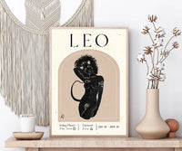 A framed art print featuring a stylized black illustration of a Leo zodiac sign on a beige background, styled on a wooden shelf next to a macrame wall hanging, a candle, and a vase with dried flowers.
