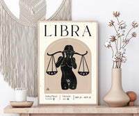 A framed Libra astrology poster displayed on a wooden shelf, styled with a macrame wall hanging, a candle, and a vase of dried cotton stems.