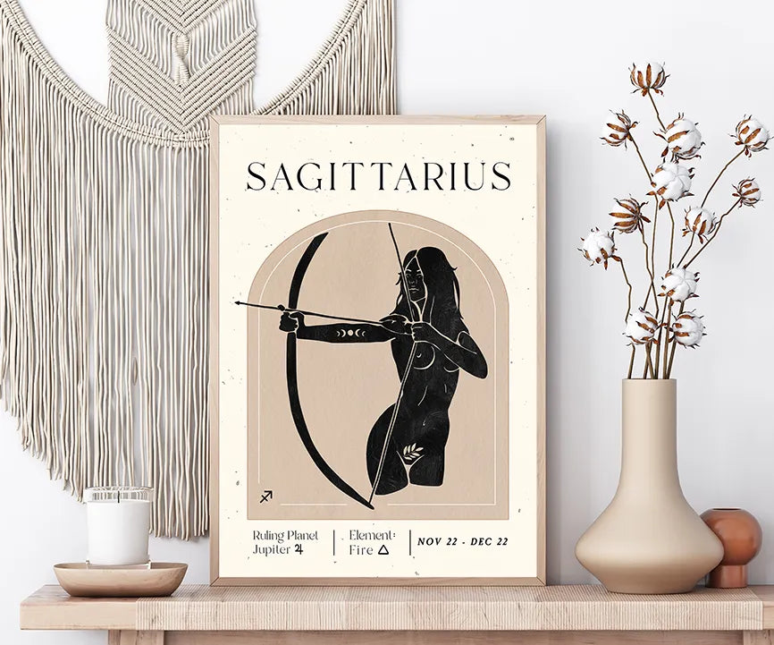A framed Sagittarius astrology poster displayed on a wooden shelf in a styled room setting, flanked by a beige macrame wall hanging, a lit candle, and a ceramic vase with dried cotton stems.