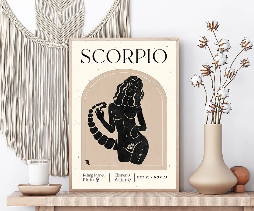 A framed Scorpio zodiac art print displayed on a wooden shelf, styled with a macrame wall hanging, a candle, cotton stems in a vase, and decorative spheres against a white wall.