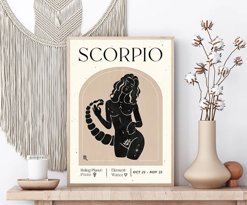 A framed Scorpio zodiac art print displayed on a wooden shelf, styled with a macrame wall hanging, a candle, cotton stems in a vase, and decorative spheres against a white wall.